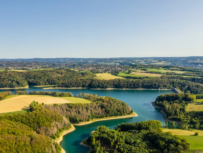 Wahnbachtalsperre Luftaufnahme eines malerischen Sees inmitten sattgrüner Wälder und Felder unter klarem Himmel.