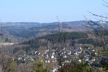 Hohe Hardt Dorf Morsbach in hügeliger Landschaft mit bewaldeten Hügeln und Windrädern im Hintergrund.