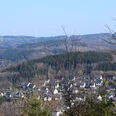 Hohe Hardt Dorf Morsbach in hügeliger Landschaft mit bewaldeten Hügeln und Windrädern im Hintergrund.