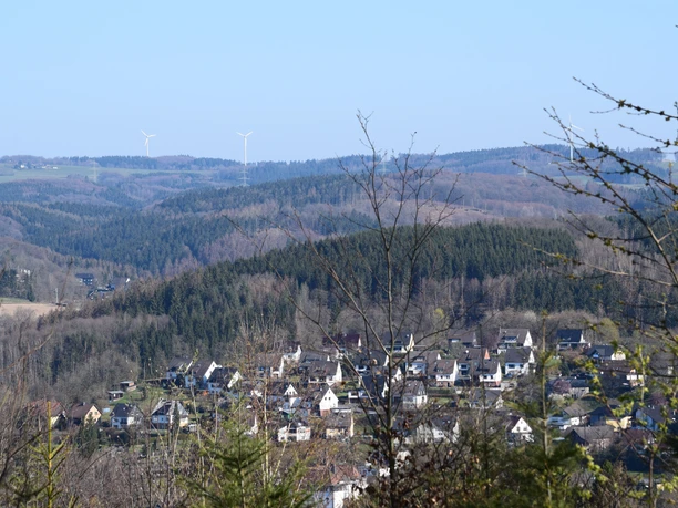 Hohe Hardt Dorf Morsbach in hügeliger Landschaft mit bewaldeten Hügeln und Windrädern im Hintergrund.