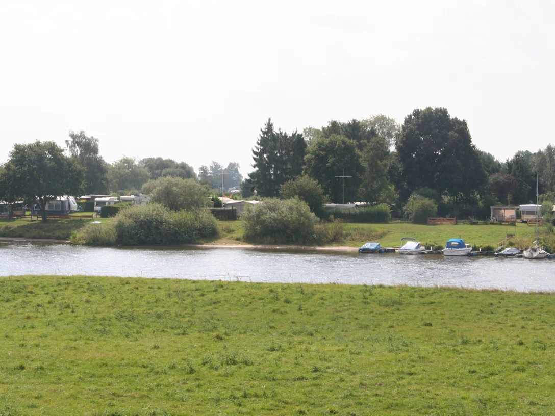 Ein grüner Campingplatz an der Weser mit Wohnwagen, Booten und Bäumen unter bewölktem Himmel.