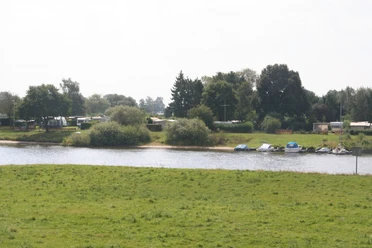 Campingplatz Fährhaus an der Weser in Thedinghausen Ein grüner Campingplatz an der Weser mit Wohnwagen, Booten und Bäumen unter bewölktem Himmel.