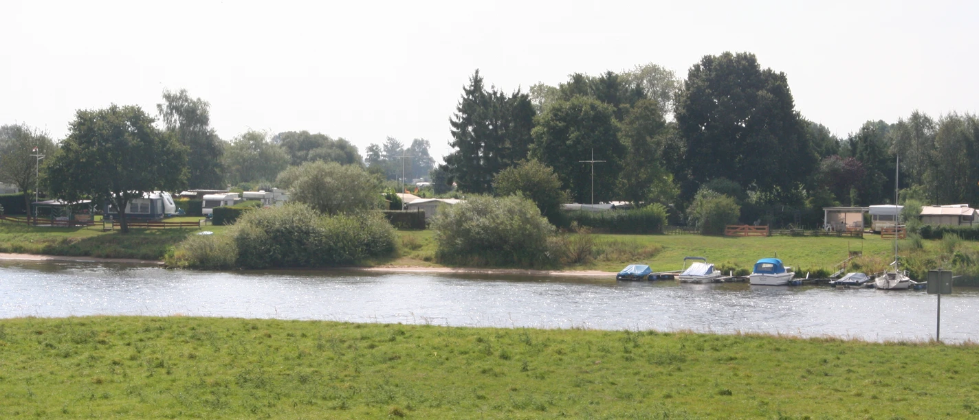 Campingplatz Fährhaus an der Weser in Thedinghausen Ein grüner Campingplatz an der Weser mit Wohnwagen, Booten und Bäumen unter bewölktem Himmel.