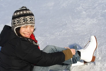Schlittschuhlaufen Eine lächelnde Frau zieht Schlittschuhe an, sitzt auf einer eisigen Fläche, bekleidet mit Winterkleidung.