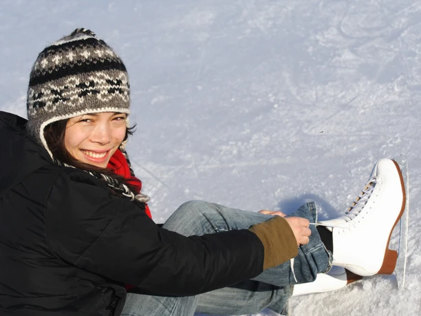 Schlittschuhlaufen Eine lächelnde Frau zieht Schlittschuhe an, sitzt auf einer eisigen Fläche, bekleidet mit Winterkleidung.