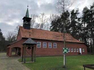 Backsteinkapelle mit grünem Kirchturm, umgeben von Rasen und Bäumen, unter bewölktem Himmel.