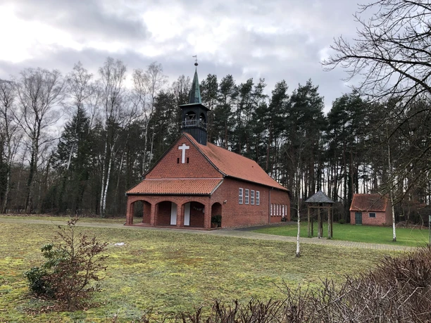 Kapelle Lichtenhorst Backsteinkapelle mit weißem Kreuz und Turm, von Bäumen umgeben, auf grünem Rasen vor bewölktem Himmel.