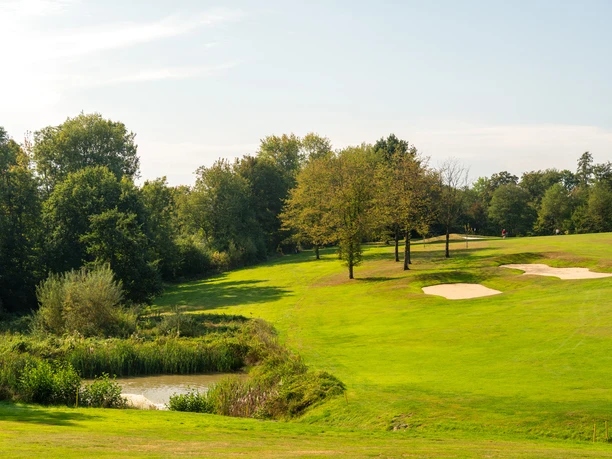 Golfplatz Burg Overbach Eine sanfte Golfplatzlandschaft mit Teich, Bäumen und Sandbunkern unter blauem Himmel.