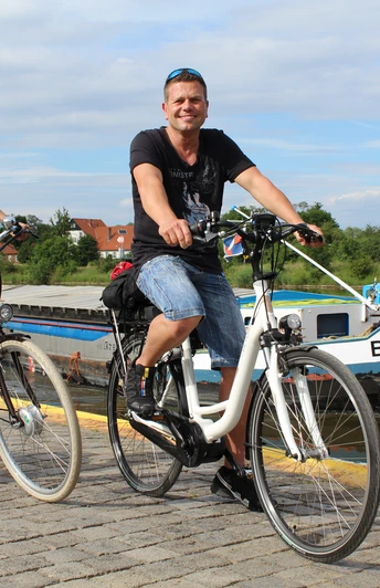 Zwei Menschen fahren auf Fahrrädern einen gepflasterten Weg entlang eines Flusses mit Boot im Hintergrund.