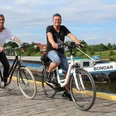 Löschplatz Stolzenau Zwei Menschen fahren auf Fahrrädern einen gepflasterten Weg entlang eines Flusses mit Boot im Hintergrund.