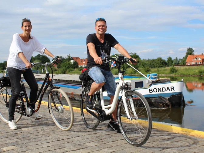 Löschplatz Stolzenau Zwei Menschen fahren auf Fahrrädern einen gepflasterten Weg entlang eines Flusses mit Boot im Hintergrund.Two people on bicycles ride along a paved path along a river with a boat in the background.To personer på cykler kører ad en brostensbelagt sti langs en flod med en båd i baggrunden.Twee mensen op fietsen rijden over een geplaveid pad langs een rivier met een boot op de achtergrond.