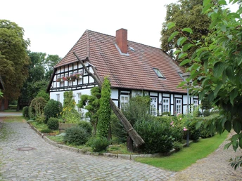 Half-timbered house with draw well in front of a well-kept garden, surrounded by trees on a paved path.