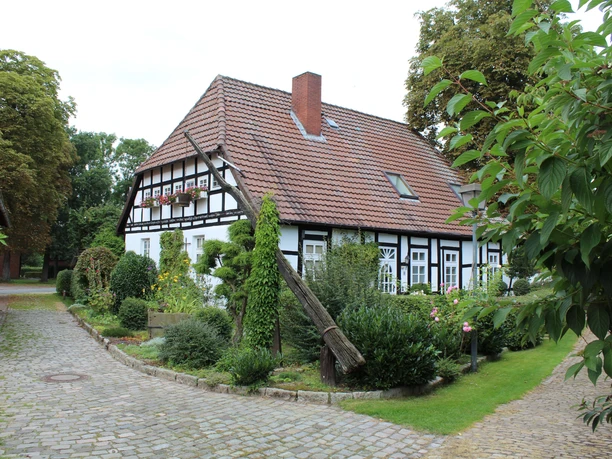 Half-timbered house with draw well in front of a well-kept garden, surrounded by trees on a paved path.