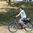Eine Person mit Helm und Sonnenbrille fährt bei Sonnenschein auf einem Elektrorad entlang einer grasbewachsenen Strecke.