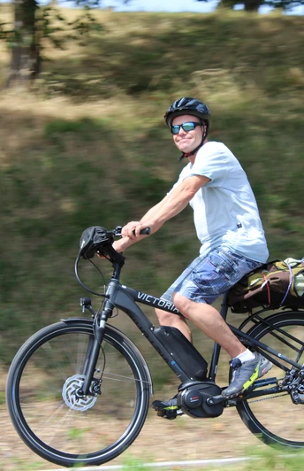 Eine Person mit Helm und Sonnenbrille fährt bei Sonnenschein auf einem Elektrorad entlang einer grasbewachsenen Strecke.