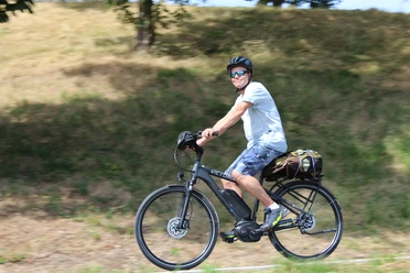 Radler Eine Person mit Helm und Sonnenbrille fährt bei Sonnenschein auf einem Elektrorad entlang einer grasbewachsenen Strecke.