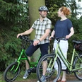 Zwei Menschen auf E-Bikes im Wald, lächelnd, bei sonnigem Wetter auf einem ruhigen Weg.