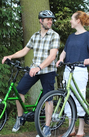 Zwei Menschen auf E-Bikes im Wald, lächelnd, bei sonnigem Wetter auf einem ruhigen Weg.