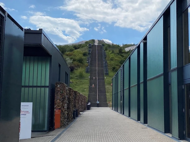 :metabolon.jpg Blick auf lange, steile Treppe zwischen modernen Gebäuden mit grünlichen Glasfassaden, Himmel teils bewölkt.