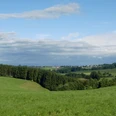 Ausblick von Hohkeppel.JPG Sanfte grüne Hügel mit Waldstreifen und verstreuten Dörfern unter blauem Himmel mit Wolkenformationen.