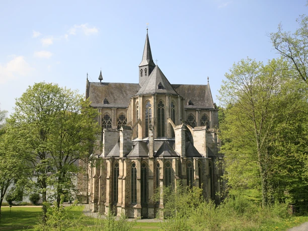 Ostchor Altenberger Dom Gotische Kirche umgeben von grünem Park mit blühenden Bäumen, blauer Himmel im Hintergrund.