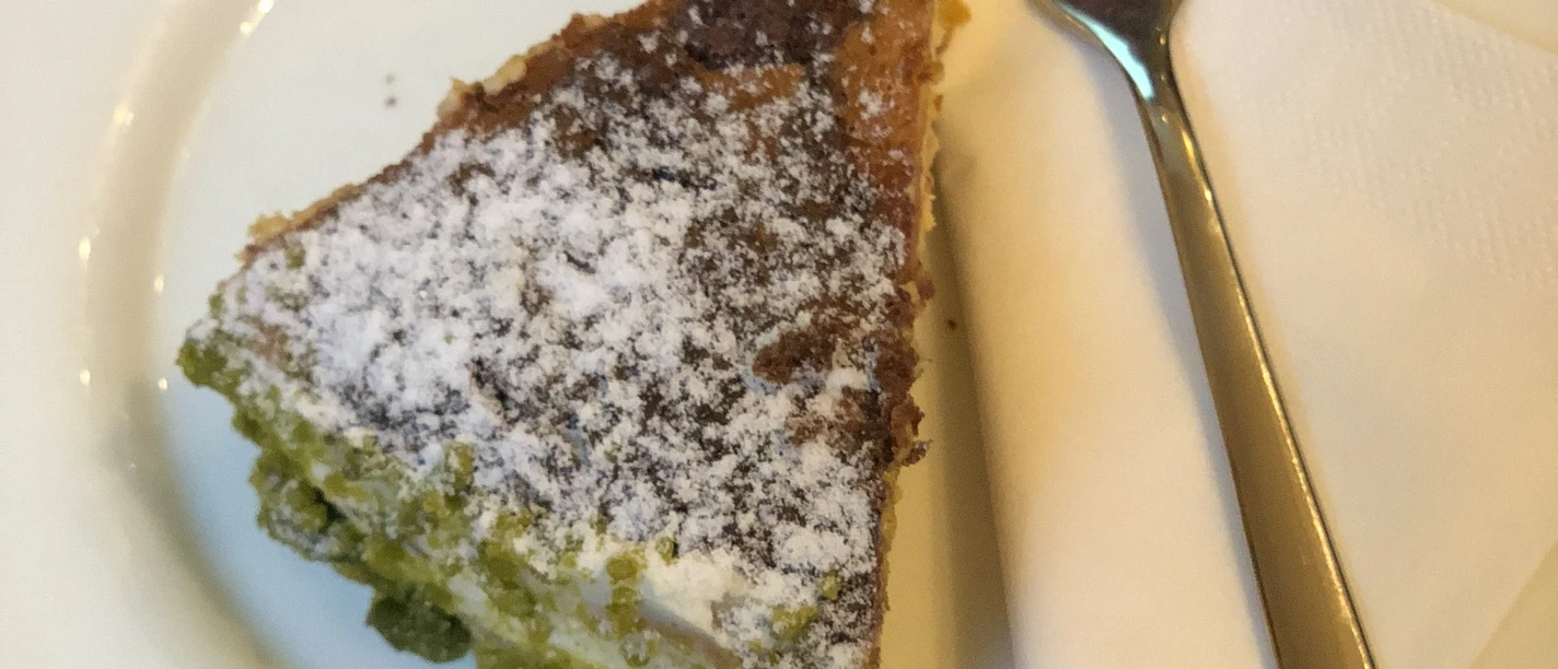 Een plak cake met groene vulling en poedersuiker, geserveerd op een wit bord met een vork.