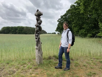 Löns Wanderweg Ein Mann steht glücklich neben einer kunstvoll geschwungenen Holzskulptur auf einem Feldweg.A man stands happily next to an artistically curved wooden sculpture on a country lane.En mand står glad ved siden af en kunstnerisk buet træskulptur på en landevej.Een man staat gelukkig naast een artistiek gebogen houten sculptuur op een landweggetje.