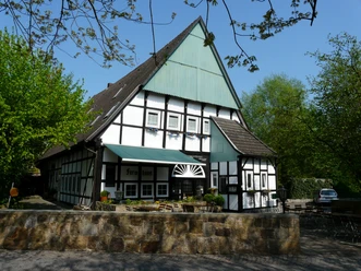 Altes Farmhaus Lienen_Frontansicht_Foto Dierkschneider.jpg Fachwerkhaus in Lienen mit grüner Markise, umgeben von Bäumen und einem gepflasterten Vorplatz.