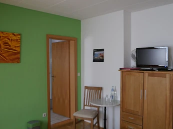 Zimmer mit grün-weißen Wänden, Holzschrank, kleiner Tisch mit Stühlen und einem Fernseher.