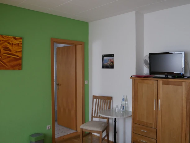 Altes Farmhaus Lienen_Zimmer_2000.jpg Zimmer mit grün-weißen Wänden, Holzschrank, kleiner Tisch mit Stühlen und einem Fernseher.
