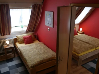 Schlafzimmer mit zwei Einzelbetten, roten Akzentwänden und Fenstern, die natürliches Licht hereinlassen.