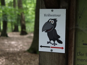 Ein Wegweiser im Wald mit einer lächelnden Krähenillustration und dem Schriftzug "Krähentour".