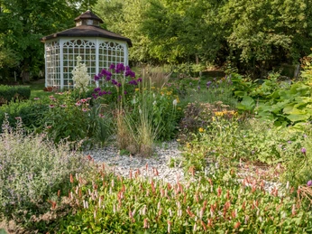 Dichte Blumenbeete mit bunten Blüten vor einem eleganten Pavillon umgeben von üppigem Grün.