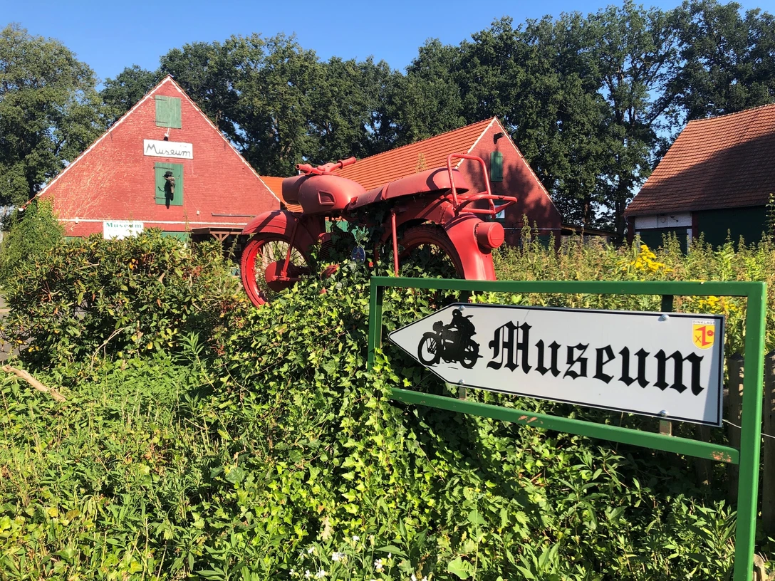 Motorrad- und Puppenmuseum Ein rotes Motorrad steht auf einem grünen Busch vor einem ziegelroten Gebäude mit Museumsschildern.