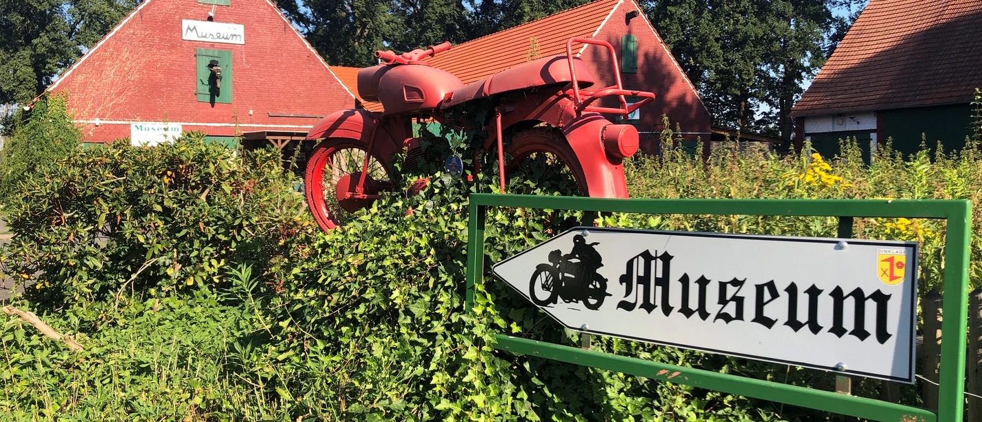 Motorrad- und Puppenmuseum Ein rotes Motorrad steht auf einem grünen Busch vor einem ziegelroten Gebäude mit Museumsschildern.