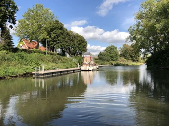 Haus, Bäume und Boot am Wasserlauf unter blauem Himmel mit weißen Wolken in Stolzenau.