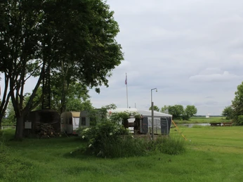 Ein grüner Rasen mit Wohnwagen und Pavillon, umgeben von Bäumen, im Hintergrund ein Fluss.