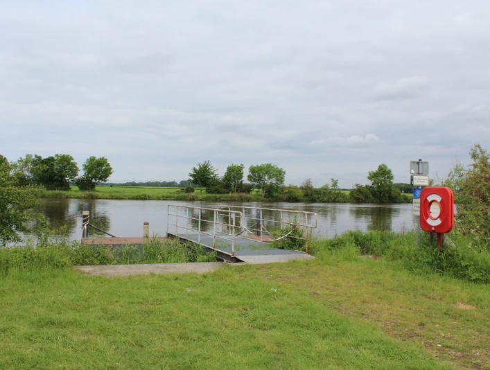 Ein Schiffsanleger an einem ruhigen Flussufer, umgeben von grüner Landschaft und bewölktem Himmel.