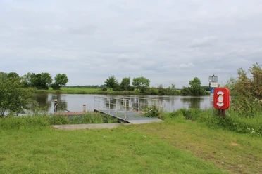 Landesbergen Schiffsanleger Ein Schiffsanleger an einem ruhigen Flussufer, umgeben von grüner Landschaft und bewölktem Himmel.