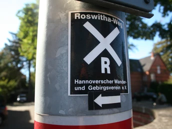 Markierung des Roswithawegs an einem Pfosten mit klar erkennbaren Symbolen und Richtungspfeil.
