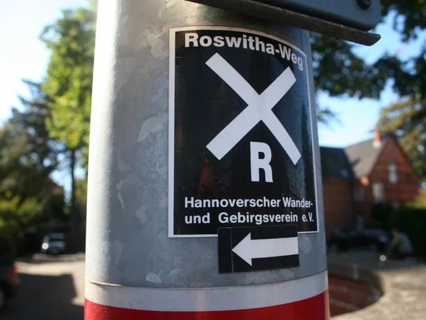 Markierung des Roswithawegs an einem Pfosten mit klar erkennbaren Symbolen und Richtungspfeil.