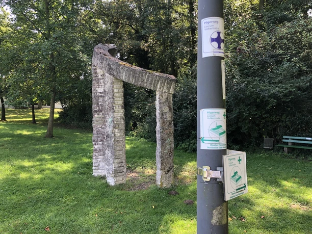 Sculpture with two arched stone steles on a green meadow next to a signpost.