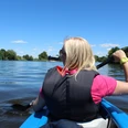 Eine Frau paddelt in einem blauen Kanu auf einem ruhigen Fluss, umgeben von grüner Landschaft.
