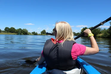 Kanutour Mittelweser Eine Frau paddelt in einem blauen Kanu auf einem ruhigen Fluss, umgeben von grüner Landschaft.