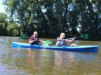 Zwei Personen paddeln in einem blauen Kanu auf einem ruhigen Fluss, umgeben von grüner Natur.