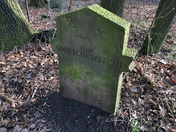 Verweerde stenen wegwijzer in het bos met "Zum Giebichenstein" erin gegraveerd, omringd door gebladerte.