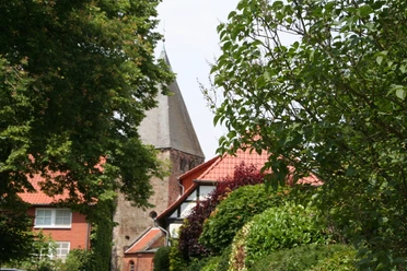 Ein Kirchturm ragt zwischen üppigen Bäumen hervor, umgeben von roten Ziegeldächern in ländlicher Kulisse.