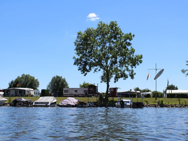 Camping grounds by the water with boat docks, trees, trailers, and a blue sky in the background.