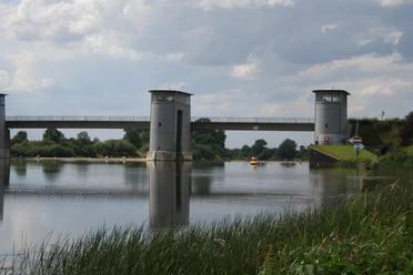 Zwei Türme eines Wehrs reflektieren sich im stillen Wasser, flankiert von grünem Ufergras.