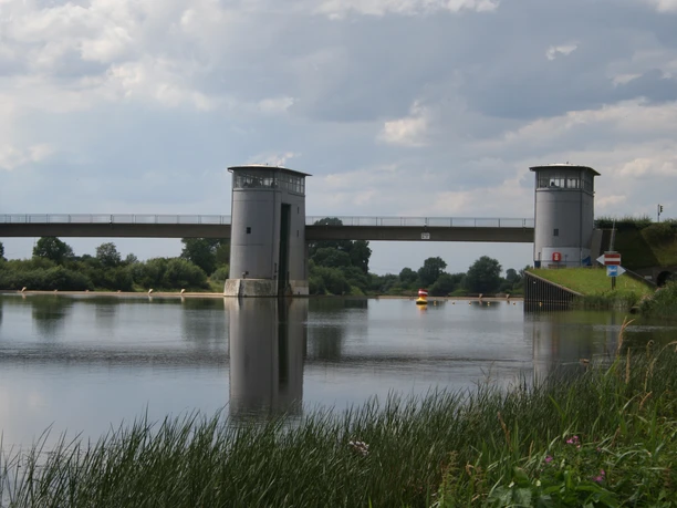Zwei Türme eines Wehrs reflektieren sich im stillen Wasser, flankiert von grünem Ufergras.